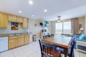 A kitchen with wooden cabinets, a dining table, and a view of the ocean through large windows. - Beachfront resort condo with Gulf views & pools/hot tub/lazy river/gym (Panama City Beach)