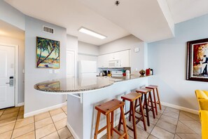 A modern kitchen with a curved island, wooden stools, and a painting of a tropical scene. - Gulf-front condo with pool, hot tub, & same-floor parking (Panama City Beach)