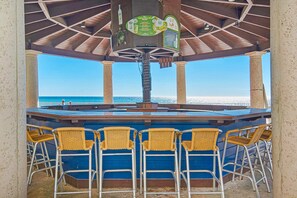 A beach bar with a view of the ocean, featuring a bar counter, wicker chairs, and a ceiling with wooden beams. - Gulf-front condo with pool, hot tub, & same-floor parking (Panama City Beach)