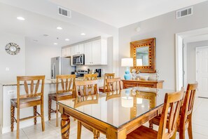 Una cocina moderna con armarios blancos, microondas y refrigerador. Hay una zona de comedor con una mesa de vidrio y sillas de madera.