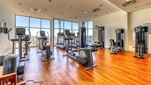 Un gimnasio amplio con diversos equipos de cardio y entrenamiento de fuerza, ventanas grandes que ofrecen vistas a la ciudad y suelo de madera.