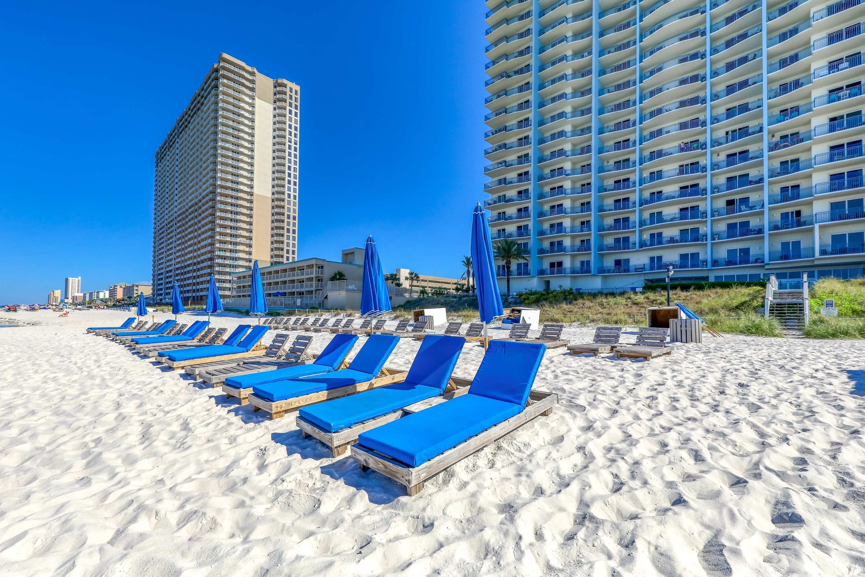 Ein Strand mit Liegestühlen und Sonnenschirmen, ein hohes Hotelgebäude sowie ein kleineres Hotel im Hintergrund.