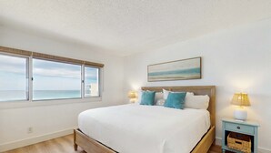 Une chambre avec un grand lit, une table de chevet avec une lampe, une fenêtre donnant sur la mer et un tableau accroché au mur.