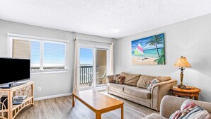 A living room with a flat-screen TV, a wooden coffee table, a sofa, and a couch. There is a painting of a beach scene on the wall. - Beautiful Pinnacle Port condo with pools & basketball court right next to beach (Panama City Beach)