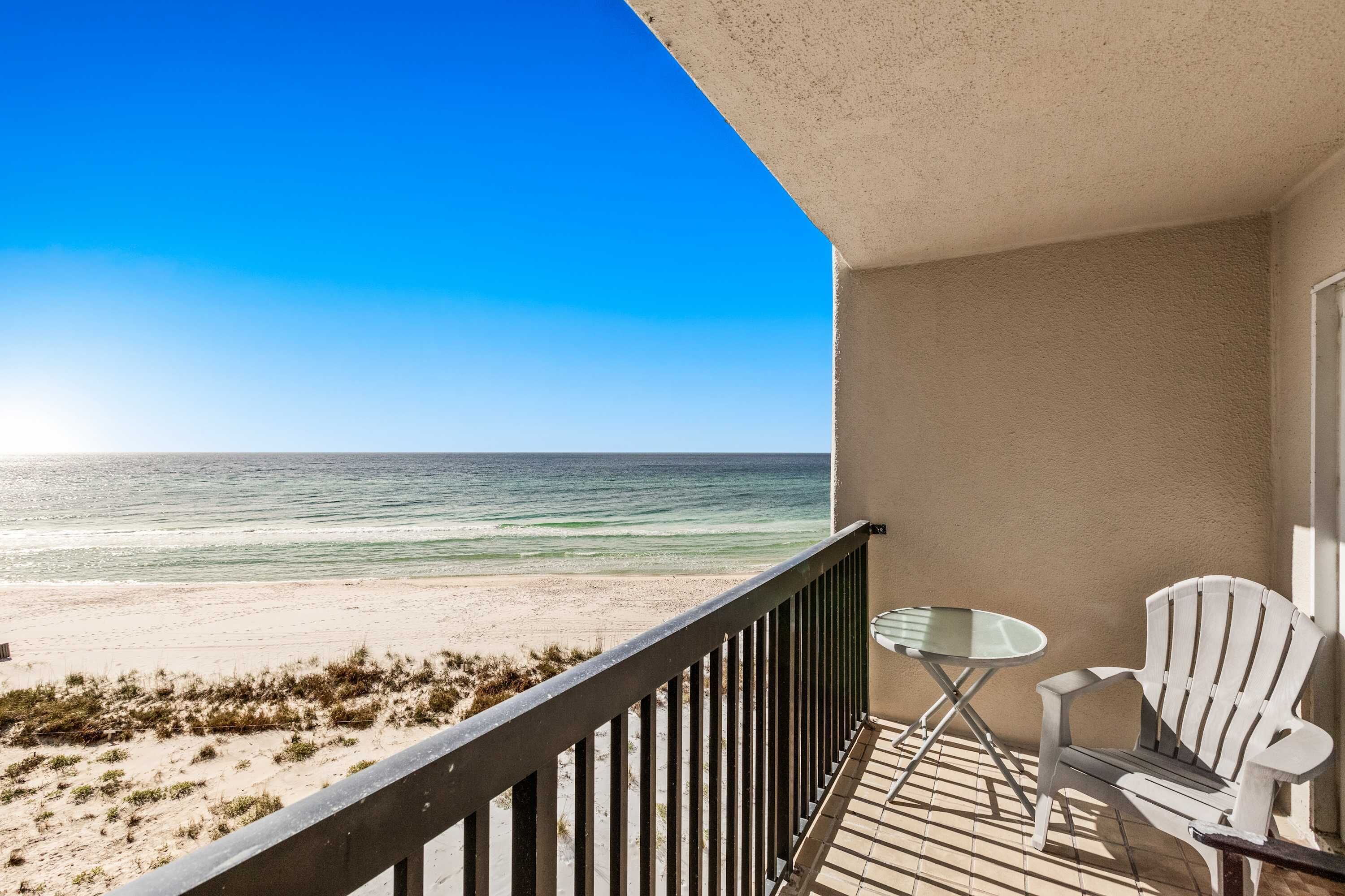 A balcony with a view of the beach, a white chair, and a small table.