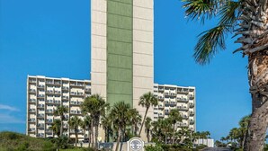 A multi-story hotel with a central green vertical feature, surrounded by palm trees and a clear blue sky. - Beautiful Pinnacle Port condo with pools & basketball court right next to beach (Panama City Beach)