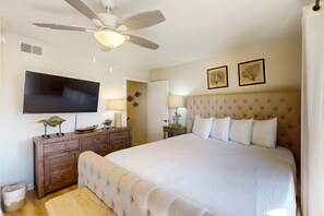 Une chambre avec un grand lit, un ventilateur de plafond, un téléviseur à écran plat, une commode en bois et deux tableaux encadrés accrochés au mur.