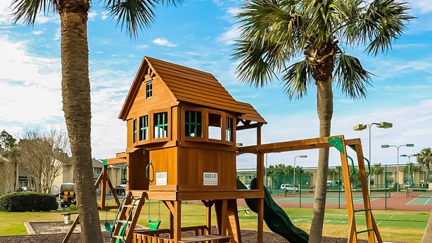 Une maison de jeu en bois, dotée d’un toboggan et de balançoires, entourée de palmiers et avec un court de tennis en arrière-plan.