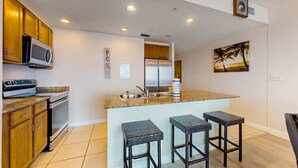 A modern kitchen with wooden cabinets, a stainless steel refrigerator, a microwave, and a granite countertop. There are three wicker barstools and a painting of palm trees on the wall. - Waterfront condo with grills, pool & fitness center - walk to the beach (Panama City Beach)