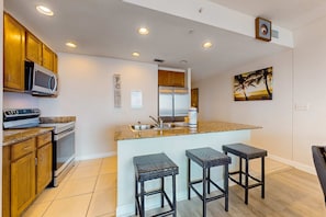 A modern kitchen with wooden cabinets, a stainless steel refrigerator, a microwave, and a granite countertop. There are three wicker barstools and a painting of palm trees on the wall. - Waterfront condo with grills, pool & fitness center - walk to the beach (Panama City Beach)