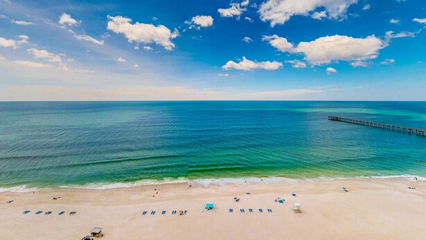 A beach with clear blue water, a pier, and beach umbrellas. - Waterfront condo with grills, pool & fitness center - walk to the beach (Panama City Beach)