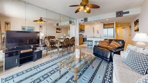 A modern living room with a TV, glass coffee table, and a sofa. There is a dining area with chairs and a table, and a kitchen in the background. - Waterfront condo with grills, pool & fitness center - walk to the beach (Panama City Beach)
