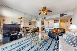 A modern living room with a TV, glass coffee table, and a sofa. There is a dining area with chairs and a table, and a kitchen in the background. - Waterfront condo with grills, pool & fitness center - walk to the beach (Panama City Beach)