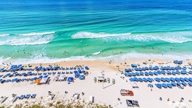 A beach with rows of blue umbrellas and sun loungers, a lifeguard tower, and a clear view of the ocean. - Beachfront, high-end condo with Pool, Hot tub & Splash pad (Panama City Beach)