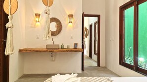 A modern bathroom with a wooden vanity, a round mirror, wall-mounted lights, and a view of a green garden through a window. - Chales em Jericoacoara by Carpediem (Jijoca de Jericoacoara)