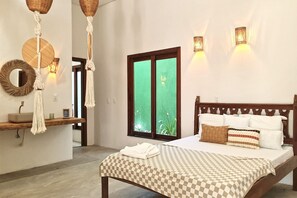 A bedroom with a wooden bed, checkered bedspread, white pillows, a wooden headboard, a bedside table, a mirror, macrame wall hangings, and a green glass door.