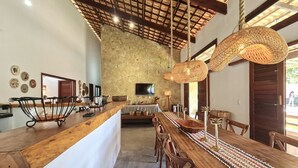 A spacious dining area with a long wooden table, wicker pendant lights, and a stone wall backdrop. - Chales em Jericoacoara by Carpediem (Jijoca de Jericoacoara)