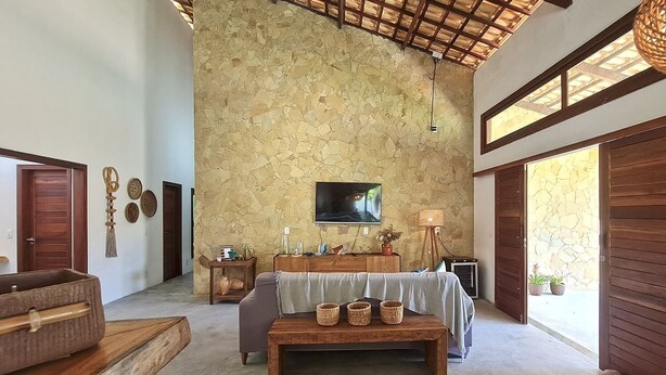 A spacious room with a stone wall, a flat-screen TV mounted above a wooden console, and a large window providing natural light. - Chales em Jericoacoara by Carpediem (Jijoca de Jericoacoara)
