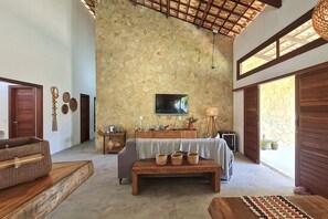 A spacious room with a stone wall, a flat-screen TV mounted above a wooden console, and a large window providing natural light.