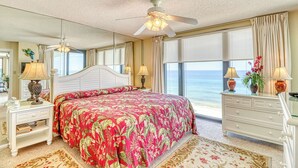 A bedroom with a large bed, a ceiling fan, a nightstand with a lamp, a dresser, and a view of the ocean. - Centrally located condo w/ private balcony, Pool & Hot tub (Panama City Beach)