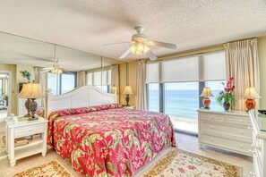 Une chambre avec un grand lit, un ventilateur de plafond, une table de chevet avec une lampe, une commode et une vue sur l’océan.