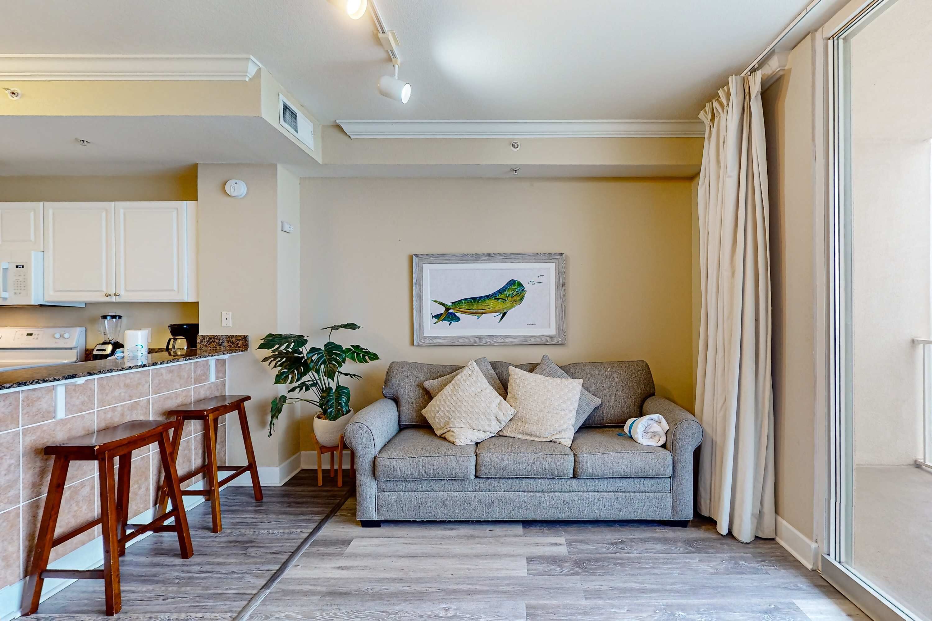 Uma sala de estar com um sofá cinzento, banquinhos de madeira, uma pintura emoldurada de um pássaro e um vaso com planta.