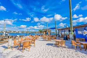 Ein Strandcafé mit Holztischen und -stühlen, einer blauen Markise und Blick aufs Meer.