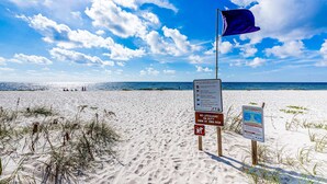 Une plage avec un ciel dégagé, un drapeau de sauveteur et des panneaux d’avertissement concernant les courants de dérive et l’absence de sauveteur.