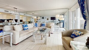 En moderne stue med sofa, sofabord og lænestol, et køkken i baggrunden og store vinduer med gardiner.
