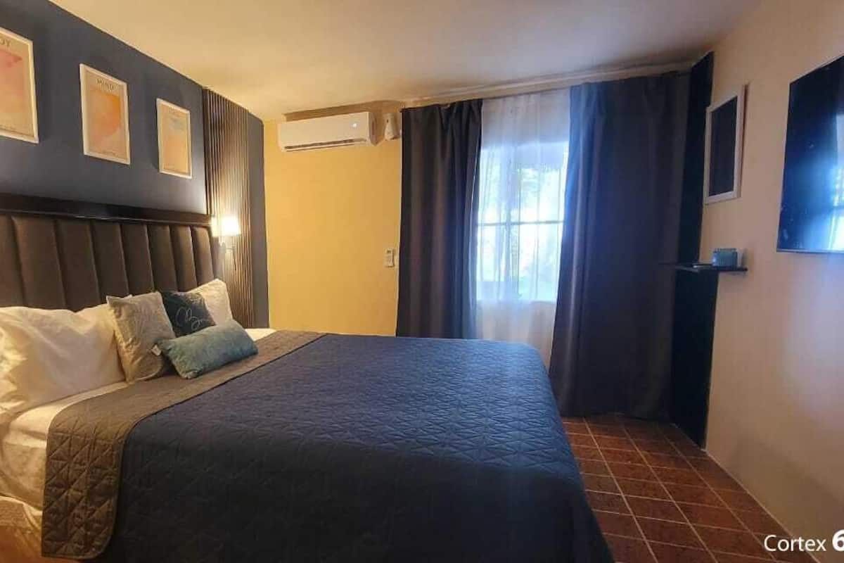 A hotel room with a large bed, a headboard, a wall-mounted air conditioner, a window with curtains, and framed pictures on the wall.