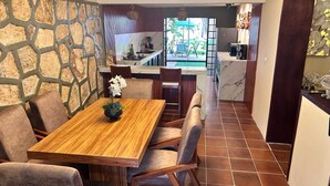 A dining area with a wooden table and chairs, a stone wall, and a view into the kitchen and outdoor seating area. - Casa Vacaional (Bacalar)
