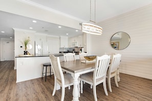 A dining area with a wooden table and white chairs, a kitchen in the background, and a hanging light fixture. - Waterfront condo with pool & hot tub access, Gulf views, W/D, & central AC (Panama City Beach)