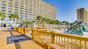 Una passerella in legno conduce a una spiaggia con un'area giochi e un grande albergo sullo sfondo.