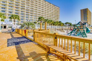 A wooden boardwalk leading to a beach area with a playground and a large hotel in the background. - Charming oceanfront condo with open layout, balcony, pool, hot tub, & gym (Panama City Beach)