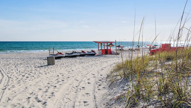 Una spiaggia con una postazione di salvataggio rossa, diversi jet ski e un sentiero di sabbia che conduce al mare.