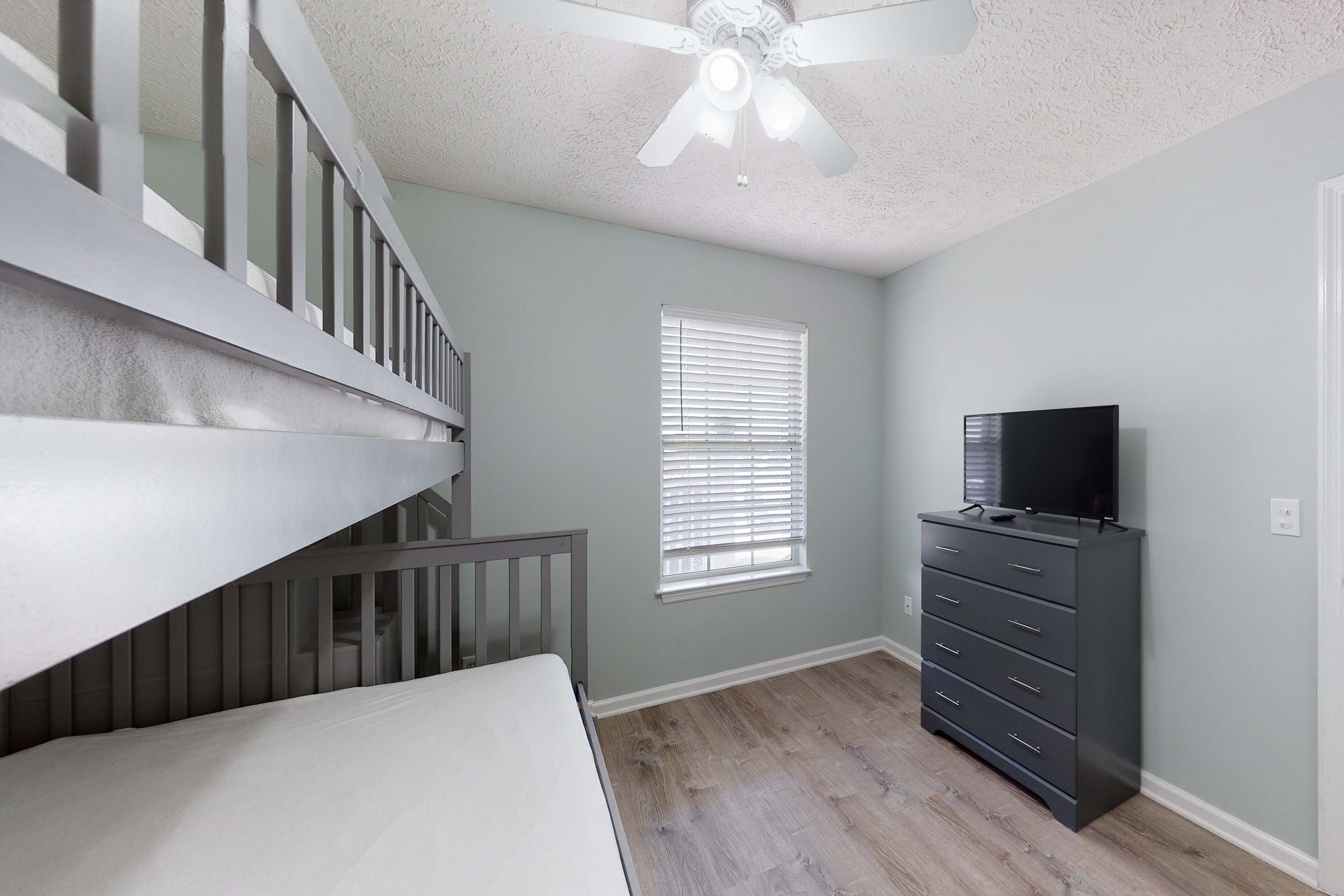Une pièce avec un escalier, un ventilateur de plafond, une fenêtre avec des stores, un téléviseur posé sur une commode et un lit.