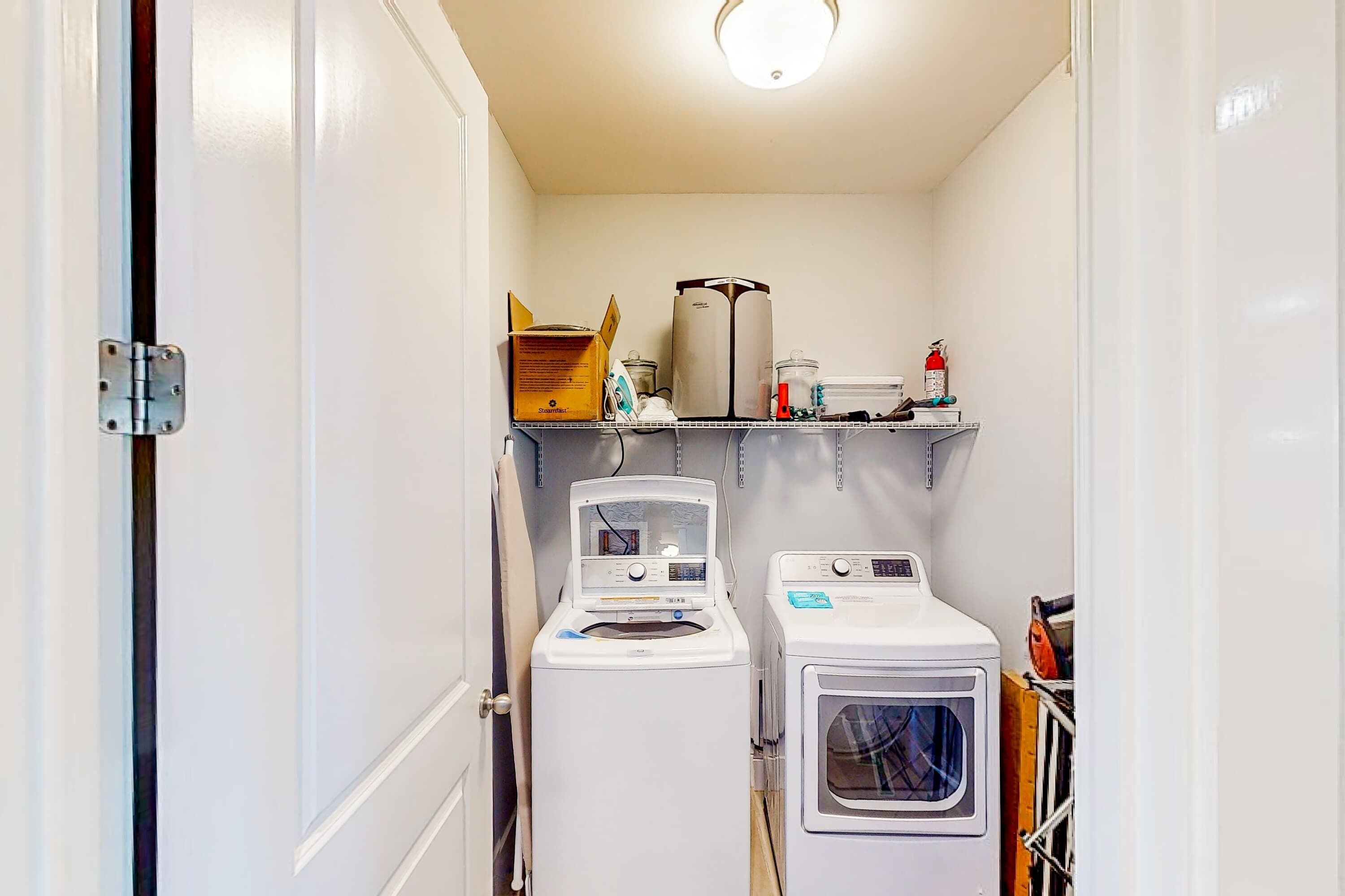 A laundry room with two washing machines and a dryer, a shelf with various items, and a door with a metal handle.