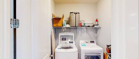 A laundry room with two washing machines and a dryer, a shelf with various items, and a door with a metal handle.