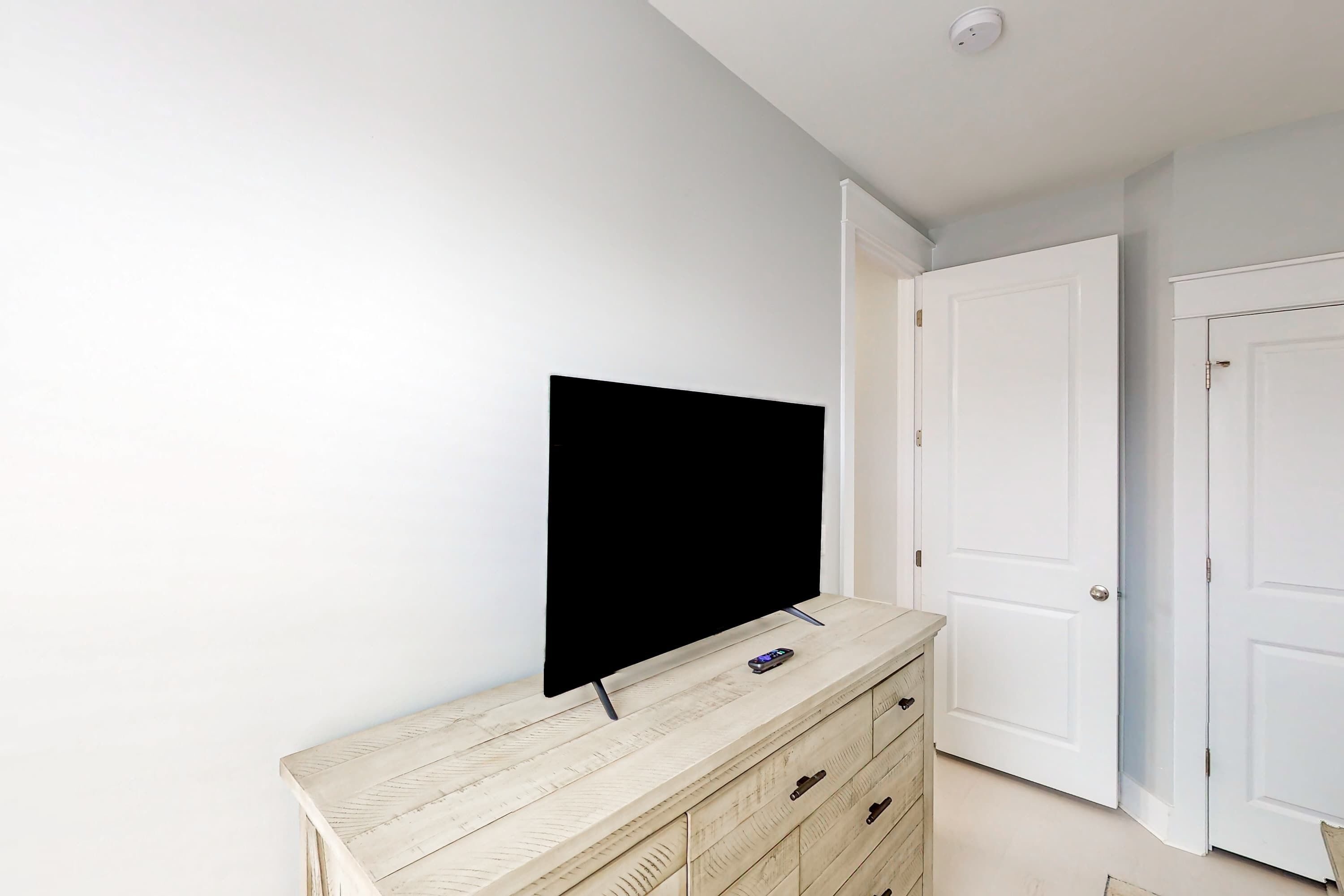 Una stanza con una TV a schermo piatto su un comò in legno, pareti bianche e una porta chiusa.
