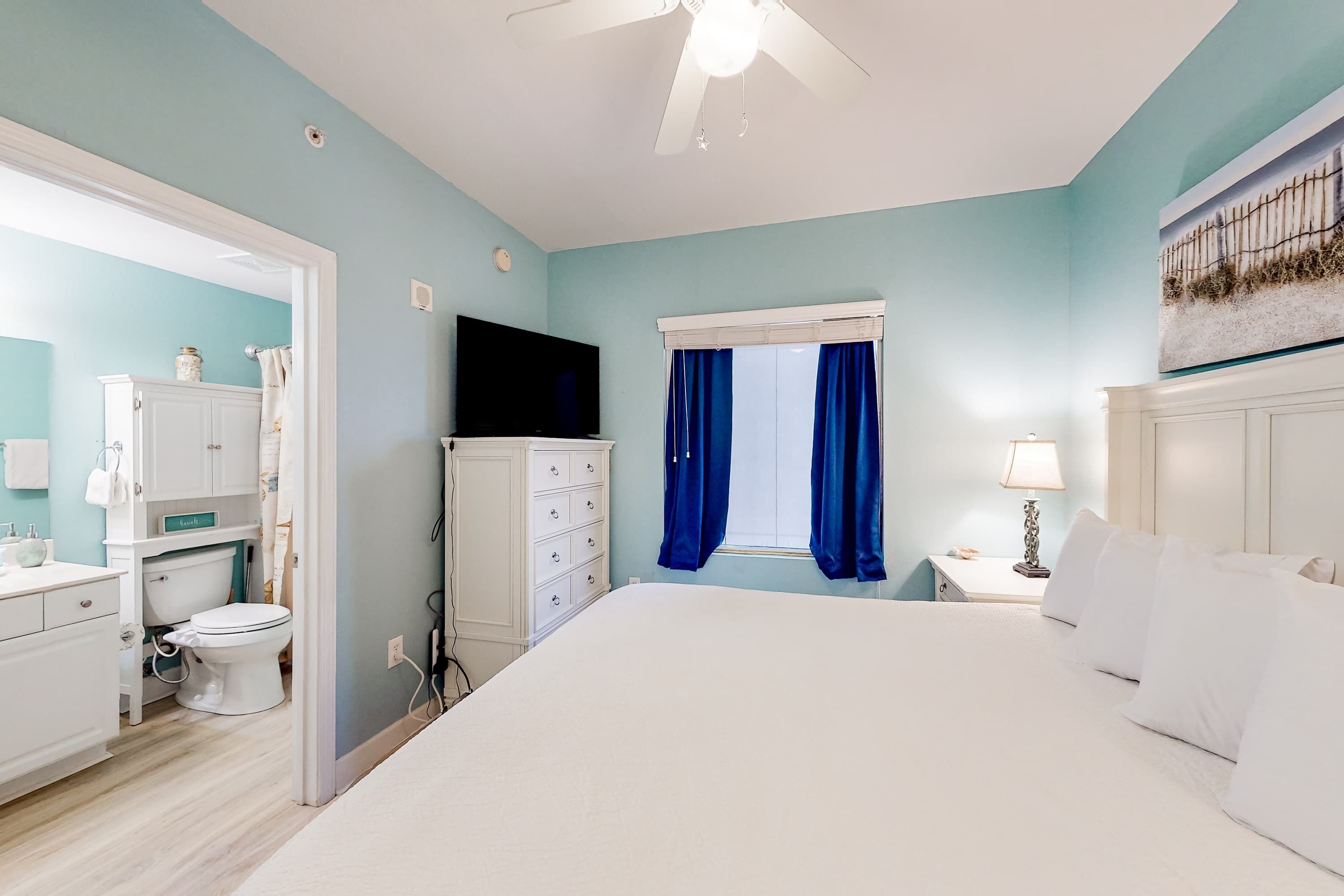 A bedroom with a bed, a TV, a window with curtains, a nightstand, and a bathroom with a toilet and cabinets.