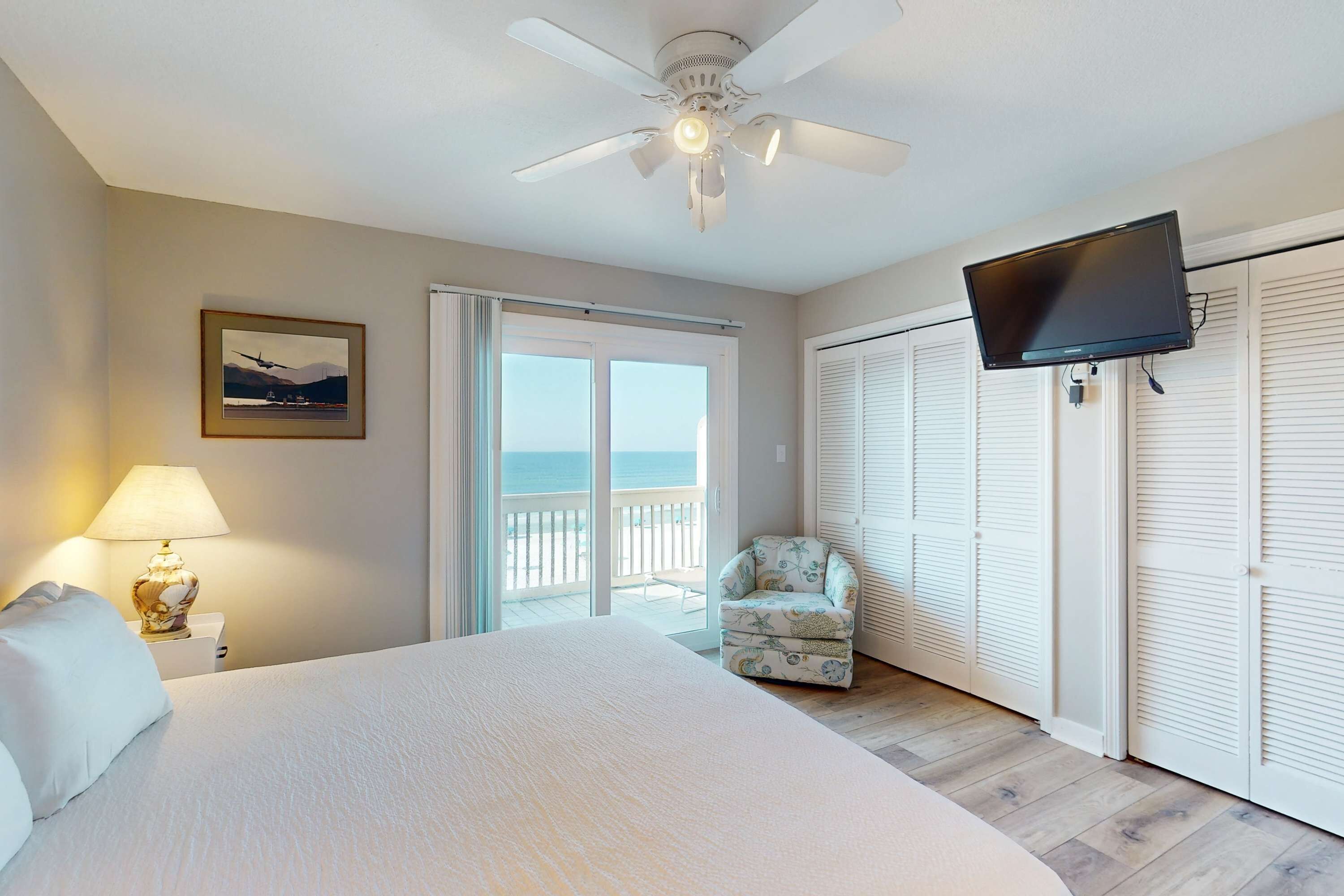 A bedroom with a bed, a ceiling fan, a TV, a chair, and a view of the ocean through a sliding door.