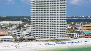 Un grand hôtel à plusieurs étages, doté d’une façade blanche et de balcons, situé près d’une plage où l’on peut voir des gens et des parasols.