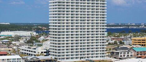 Un hotel alto de varios pisos con fachada blanca y balcones, situado cerca de una playa con gente y sombrillas de playa.