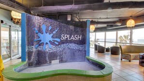 Un espace piscine agrémenté d’un mur en mosaïque sur lequel est inscrit le mot « SPLASH », entouré de sièges et doté de grandes fenêtres.