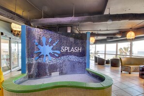 Ein Poolbereich mit einer Wand aus Mosaiken, auf der "SPLASH" steht, umgeben von Sitzgelegenheiten und großen Fenstern.