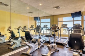 A gym with multiple exercise machines, large windows, and a view of the ocean. - Tropical-themed condo with bi-level pool, fitness center & beach access (Panama City Beach)