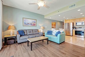 A living room with two sofas, a coffee table, and a ceiling fan. There is a kitchen area with a refrigerator, microwave, and oven. - Gulf front condo with resort amenities, snack bar, and Lazy River (Panama City Beach)
