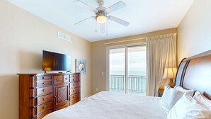 A hotel room with a large bed, a wooden dresser, a television, and a balcony with curtains. - Wonderfully decorated Gulf-view condo | Pool | Hot tub (Panama City Beach)