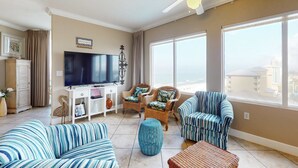 A living room with a TV, wicker chairs, a striped sofa, and large windows. - Lovely condo at beachfront resort with sunset views, pools, hot tubs, & gym (Panama City Beach)