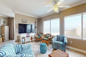 A living room with a TV, wicker chairs, a striped sofa, and large windows. - Lovely condo at beachfront resort with sunset views, pools, hot tubs, & gym (Panama City Beach)